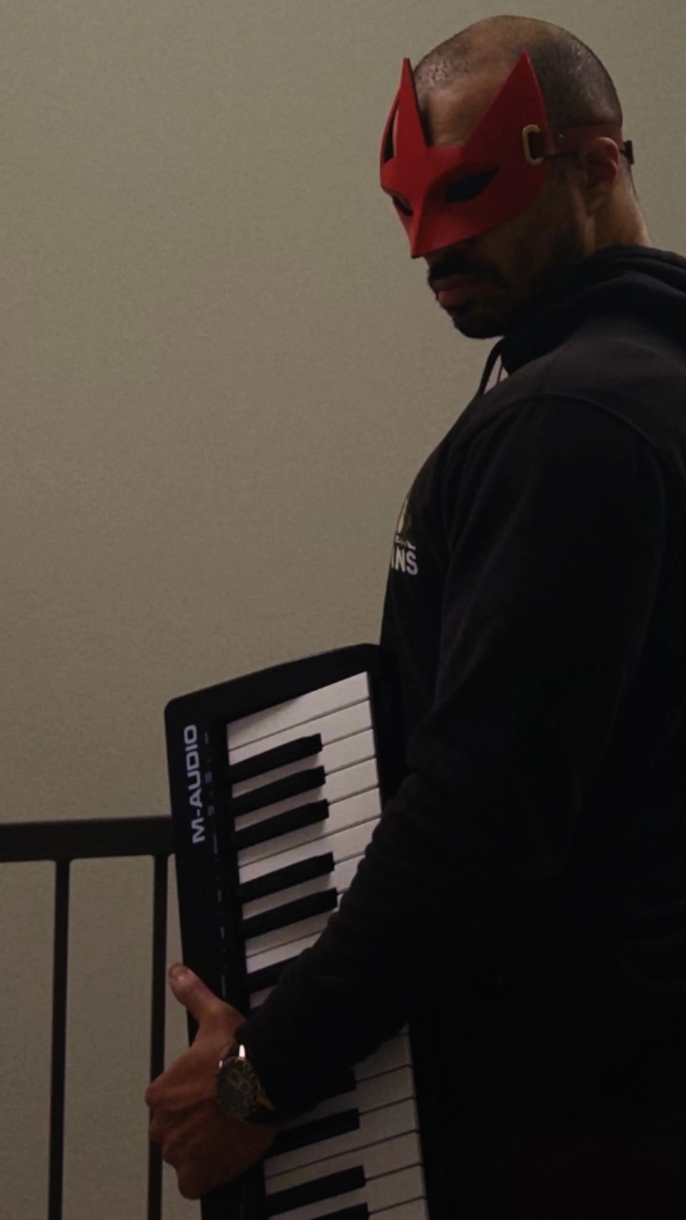 A picture of JSTNURMIND (Just In Your Mind), a red masked musician, holding a piano midi controller.