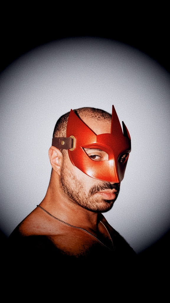 Close-up portrait of JSTNURMIND wearing the red faceted mask, lit by a soft spotlight.
